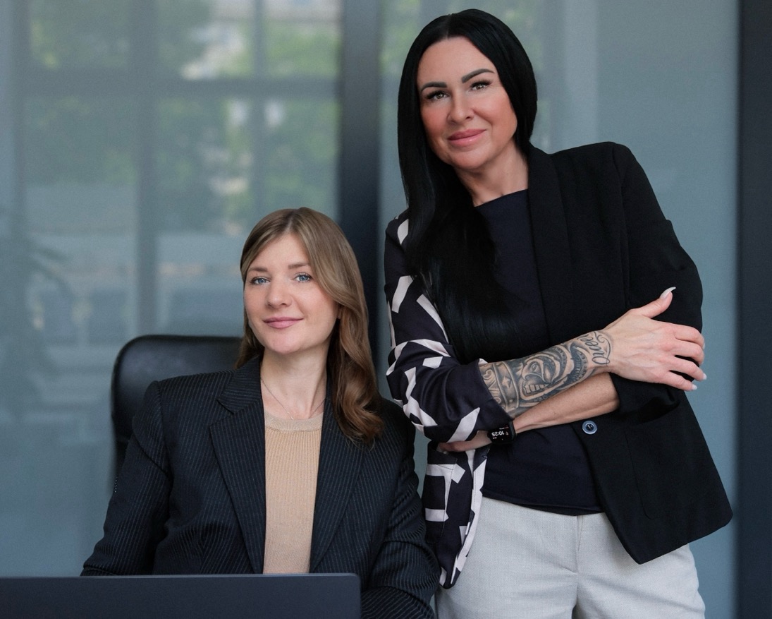 Zwei lächelnde Frauen im Büro; eine sitzt, die andere steht mit tätowiertem Arm und dunklem Haar.